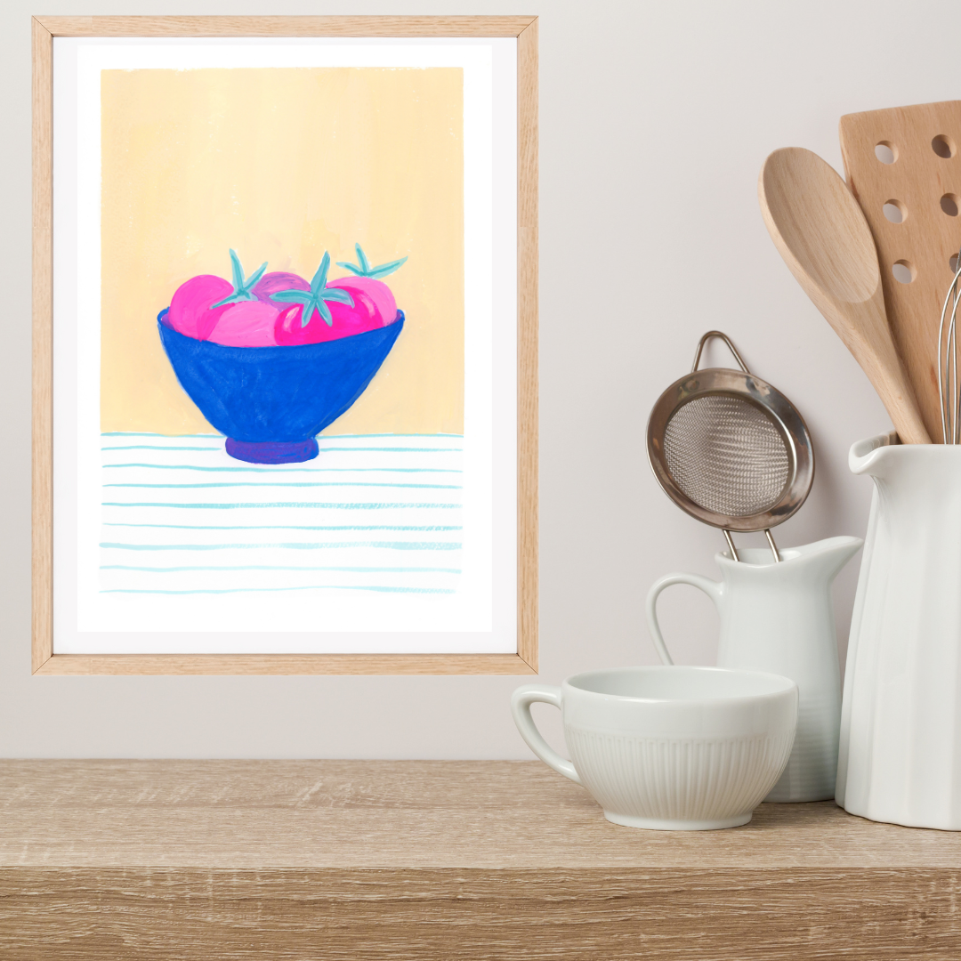 A framed original gouache painting featuring a blue bowl of pink tomatoes on a light yellow background, styled on a wooden shelf next to white ceramic kitchen items including a cup, pitcher, and wooden utensils.