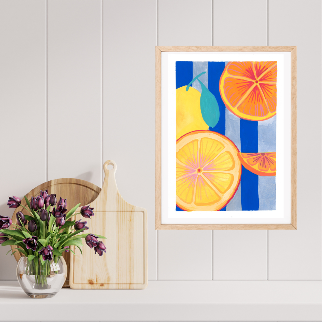 A framed original featuring a colourful gouache illustration of citrus fruits (orange slices, a yellow pear, and a green lime) on a white panels wall, styled next to a wooden cutting board and a vase of purple tulips.