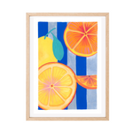 Framed wall art featuring a colourful illustration of citrus fruits, including a whole yellow lemon and several orange slices, set against blue and light gray vertical stripes. The artwork is presented in a light natural wood frame with a white mat on a plain white background.
