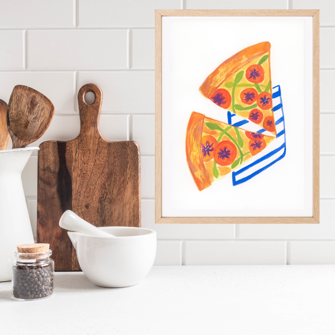 A framed art print featuring a colourful gouache illustration of two slices of pepperoni pizza on a white background, styled on a kitchen counter next to wooden cutting boards, a white bowl, and a jar of peppercorns.