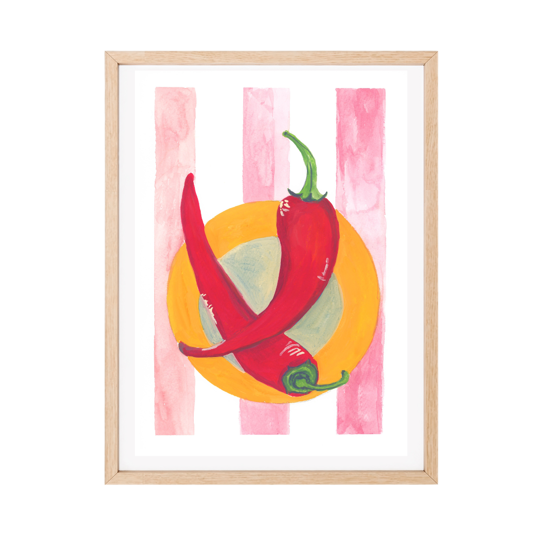 Framed original gouache artwork featuring two red chili peppers on a yellow orange plate against pink vertical stripes, in a light natural wood frame with a white mat.