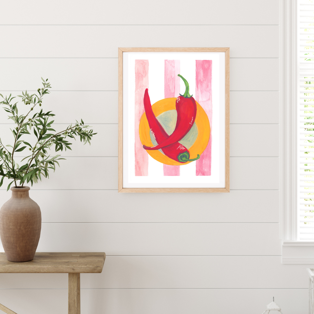 A framed original gouache artwork featuring two red chili peppers illustration in an orange plate and on pink striped background, hanging on a white shiplap wall next to a wooden table with a vase of green leaves.