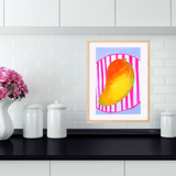 A framed original gouache artwork featuring a colourful mango on a pink striped background, displayed on a black countertop against a white subway tile backsplash.